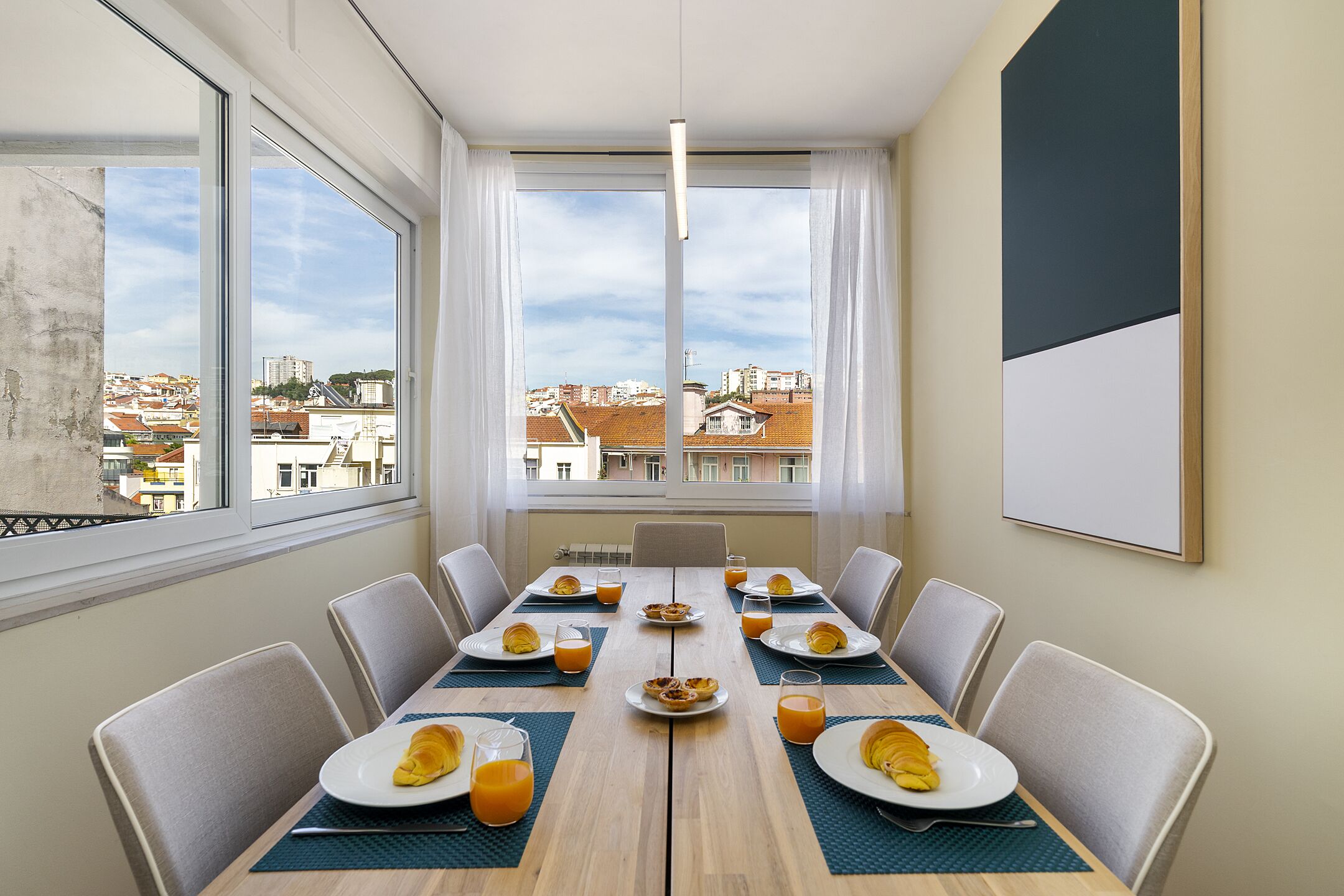 A dining area with a table set for eight, large windows with curtains, and a view of the cityscape.