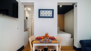 A small dining area with a table set for two, a TV mounted on the wall, and a bed in the adjacent room.