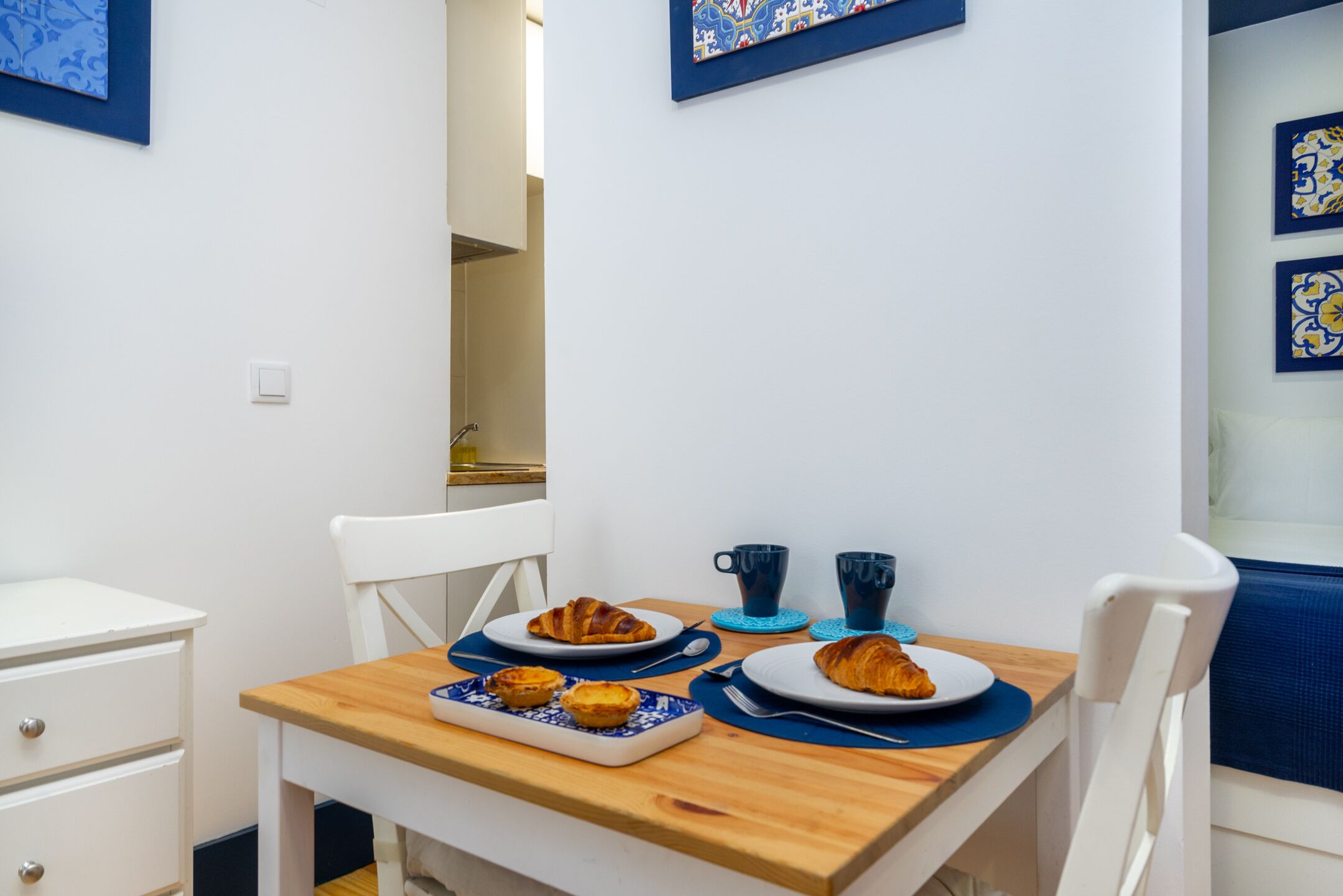 A breakfast table with pastries and coffee cups, a white chair, and a blue wall with artwork.