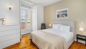 Ein Schlafzimmer mit einem Bett, Nachttischlampen, einem Sessel, einer Tür, die auf einen Balkon führt, und einem gerahmten Bild an der Wand.