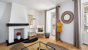 Ein Wohnzimmer mit Kamin, einem weißen Sessel, einem gelben Stuhl, einem runden Spiegel und einem Glastisch.