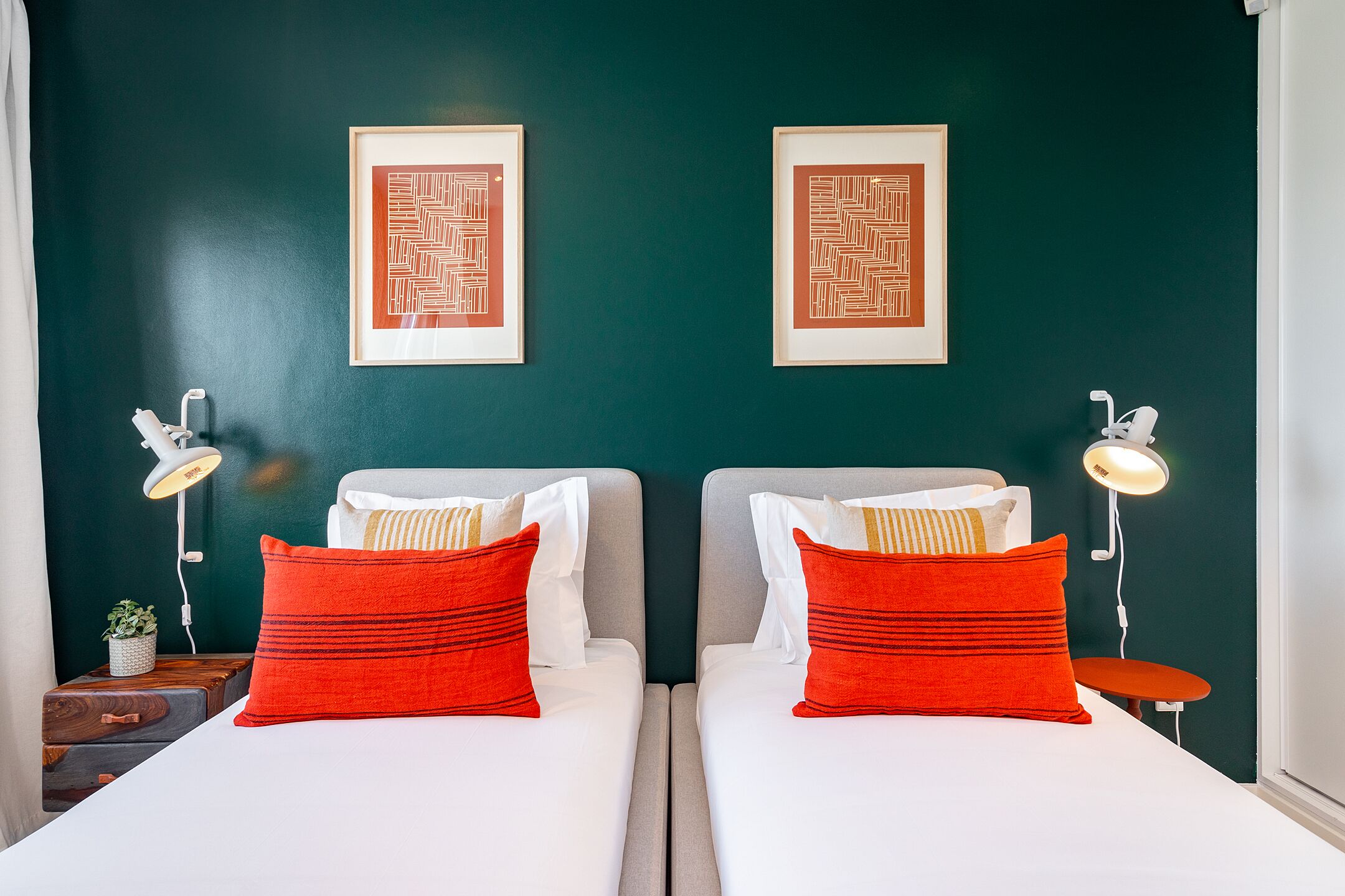 A hotel room with two beds, each with a red pillow and white bedding. There are framed artworks on the wall, and bedside lamps on either side of the beds.