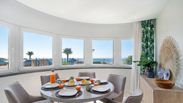 Un comedor con una mesa redonda dispuesta para el desayuno, ventanas amplias con vista al mar y un adorno en la pared.