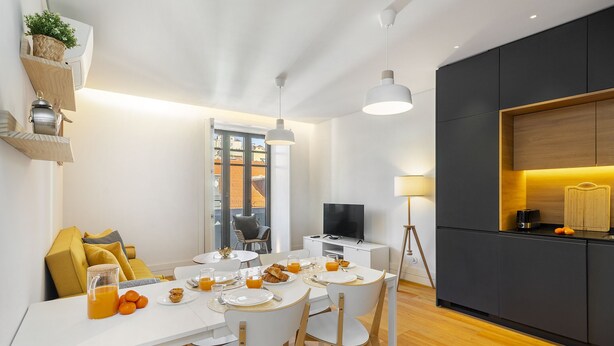 A modern dining area with a white table, wooden chairs, and a kitchen area with dark cabinets and a TV. - Urban Elegance: A Modern Haven In Lisbon'S Heart (Lisbon)