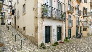 A cobblestone street with a building featuring a balcony with a wrought iron railing, a motorcycle parked outside, and a staircase leading to the entrance. - Cozy Alfama Haven: Perfect For Exploring Lisbon (Lisbon)