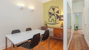 Un coin repas avec une table blanche et des chaises noires, un meuble en bois surmonté d’une lampe, et un mur orné d’un motif géométrique.