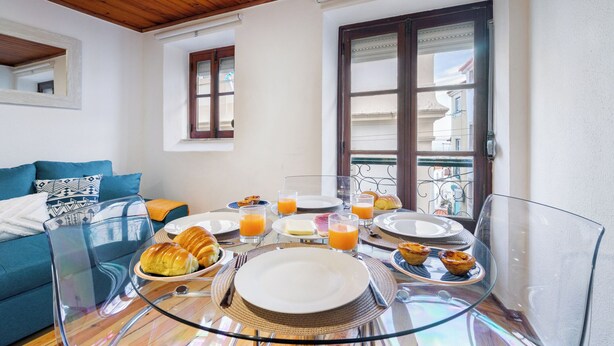 A breakfast setup with croissants, orange juice, and pastries on a glass table in a room with a wooden ceiling and a balcony view. - Beco Patacas Perfect Location To Explore Lisbon (Lisbon)