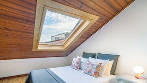 A bedroom with a sloped ceiling, a skylight, a bed with colorful pillows, a bedside table with a lamp, and a view of a building through the skylight. - Beco Patacas Perfect Location To Explore Lisbon (Lisbon)