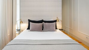 A neatly made bed with a white duvet and two pillows, flanked by two bedside tables with lamps, in a room with a wooden floor and white walls. - Luxury Stay In Prime Location W/ Ac, Lift & Private Patio (Lisbon)