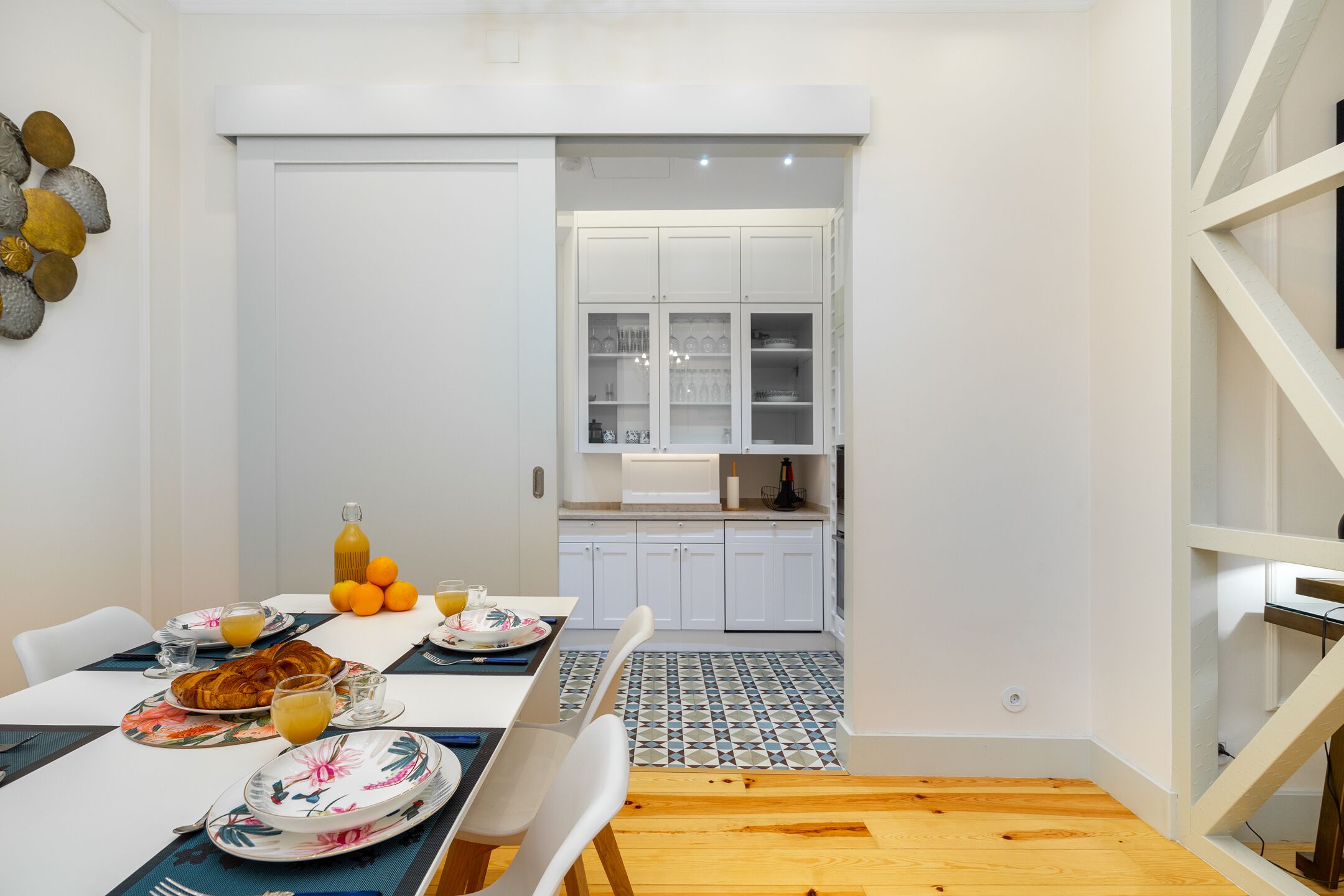 Un coin repas avec une table mise pour le petit-déjeuner, une cuisine avec des meubles blancs et un sol carrelé.