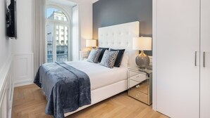Un dormitorio moderno con una cama grande, dos mesitas de noche con lámparas, un televisor montado en la pared y vistas de la ciudad a través de un ventanal amplio.