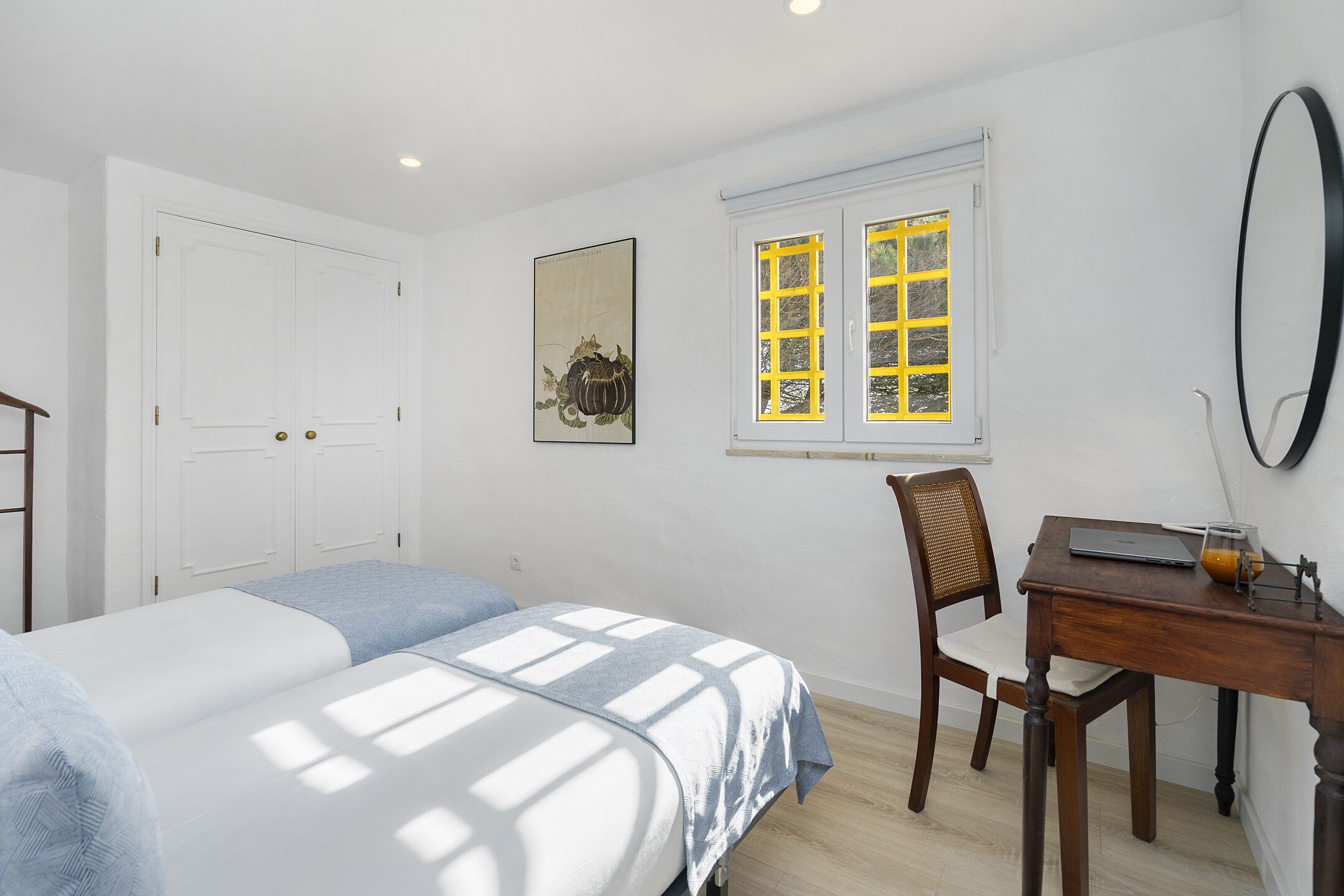 A bedroom with two beds, a desk, a chair, a window with yellow shutters, and a wall-mounted mirror.