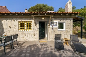 A stone building with a tiled roof, a black door, and a small window. There are two outdoor tables with chairs, one on each side of the building. - Wilderness Escape: Secluded Haven For Remote Work (Sintra)