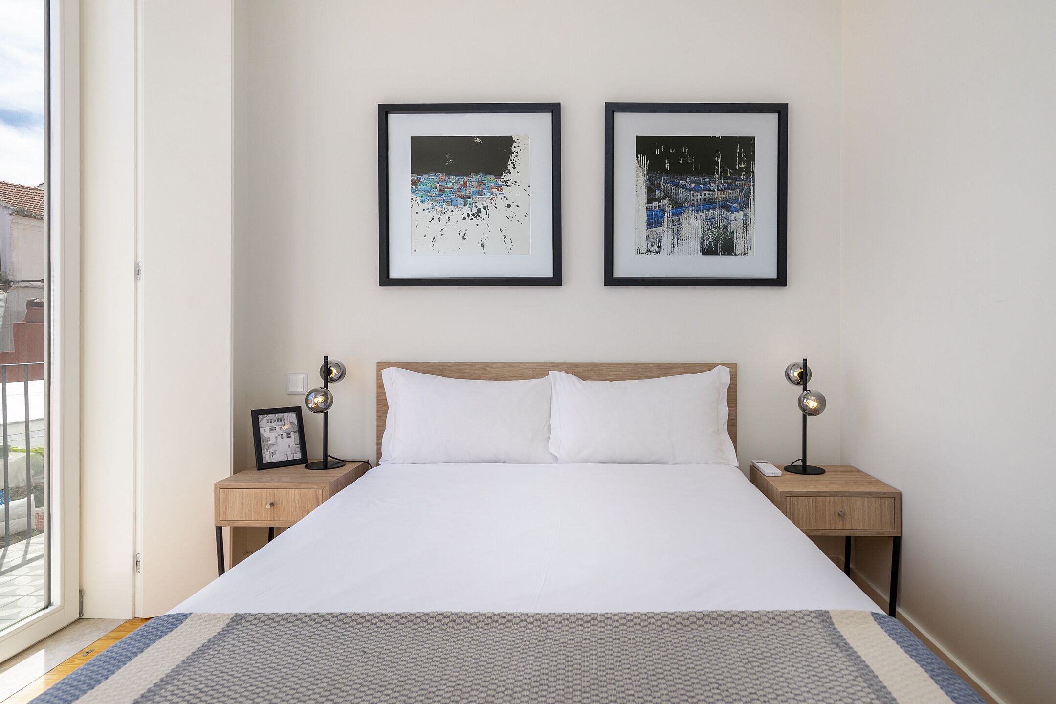 A neatly made bed with white linens, two framed artworks above it, and a nightstand with a lamp on each side.