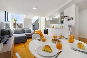 Ein modernes Wohnzimmer mit einem grauen Sofa, einem Esstisch mit Croissants und Orangensaft sowie einer Küchenzeile mit weißen Schränken und einer Spüle.