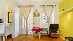 Une pièce avec un plancher en bois, une table à manger avec des chaises, un téléviseur posé sur un meuble et une fenêtre avec des rideaux.