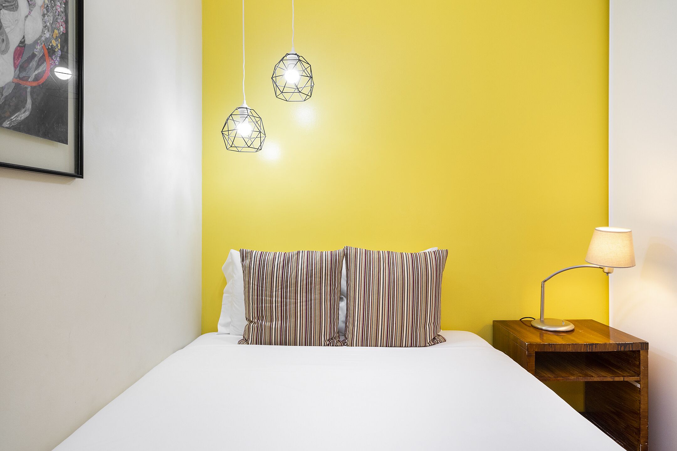 A bed with striped pillows, a bedside table with a lamp, and geometric pendant lights.