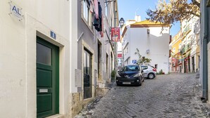Une ruelle étroite pavée de pierres, bordée de bâtiments des deux côtés, avec une porte verte et des voitures garées.