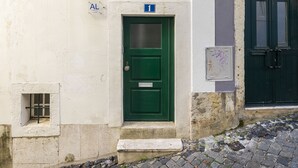 Une porte verte avec une petite fenêtre et une plaque métallique, un mur blanc avec un panneau indiquant un numéro, et une porte vert foncé avec une poignée métallique.