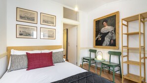 Ein ordentlich bezogenes Bett mit einer schwarzen Decke, weißen und roten Kissen sowie einem hölzernen Kopfteil. An der Wand hängen gerahmte Bilder, ein grüner Sessel mit einer Lampe und ein Holzregal.