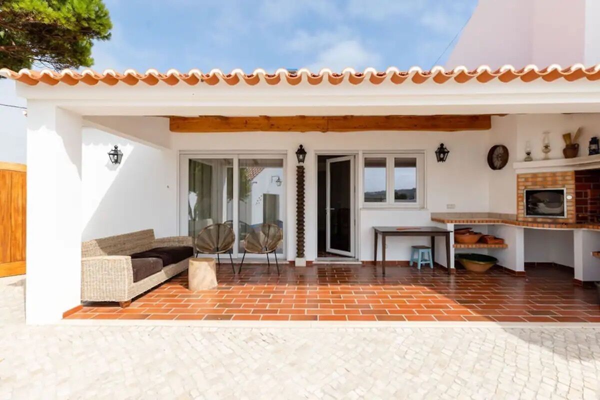 A covered patio with a brick floor, a white wall, a wooden ceiling, a sofa, two chairs, a table, and a fireplace.