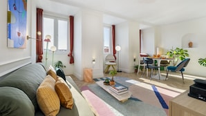 A modern living room with a sofa, a coffee table with books and a plant, a dining area with chairs and a table, and a large window with curtains. - Historic Centre Modern Masterpiece W River Views (Lisbon)