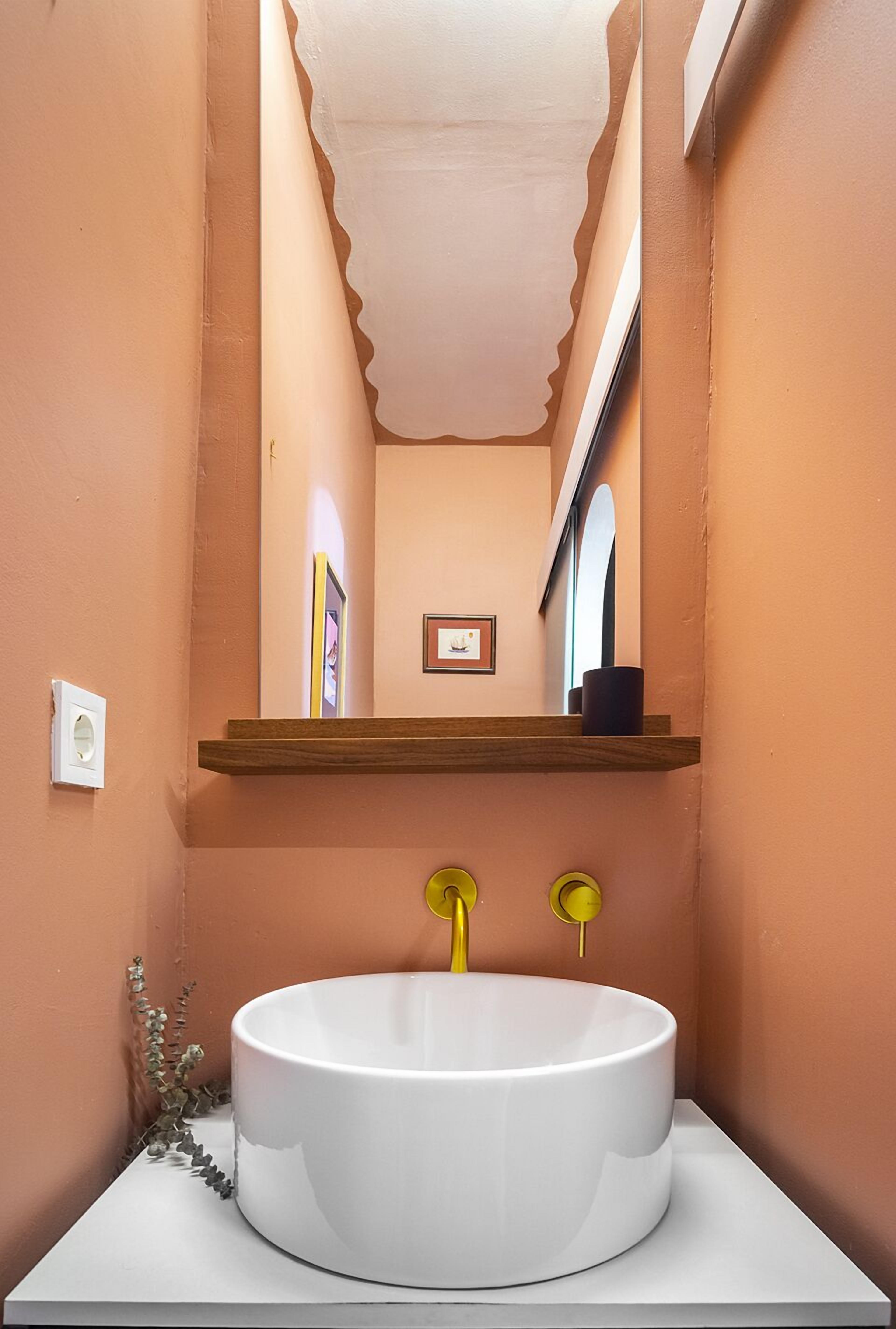A bathroom with a round white sink, yellow faucets, and a wooden shelf above the sink.