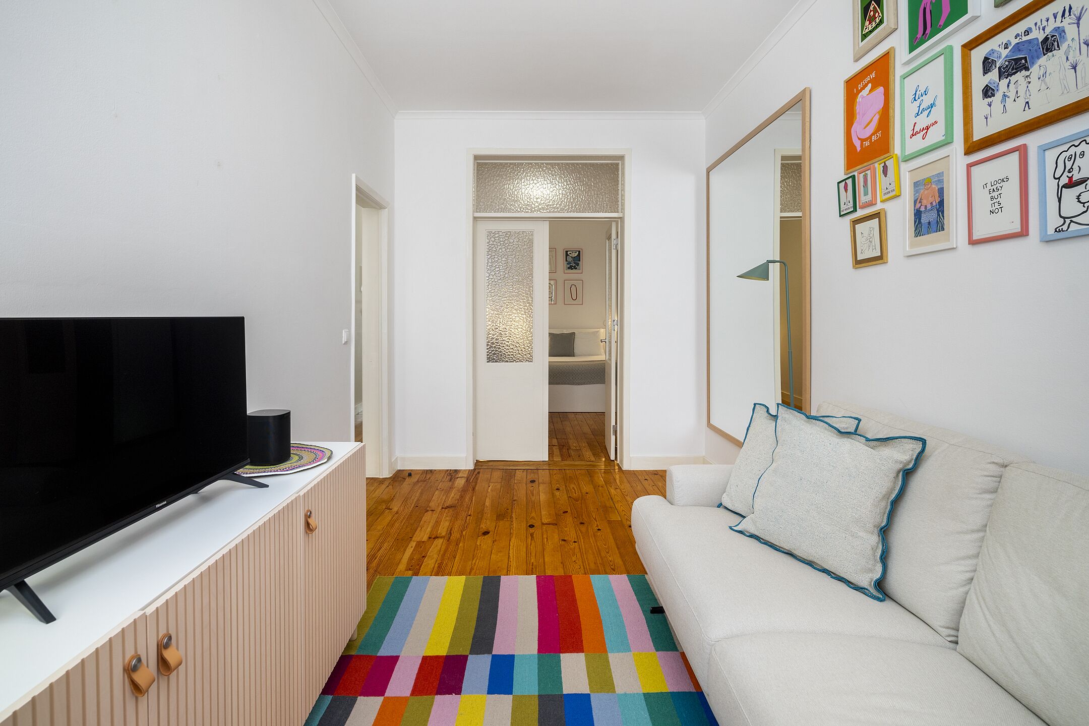 A modern living room with a flat-screen TV on a white cabinet, a colorful striped rug, a white sofa with a blue pillow, and a hallway leading to a room with a glass door.