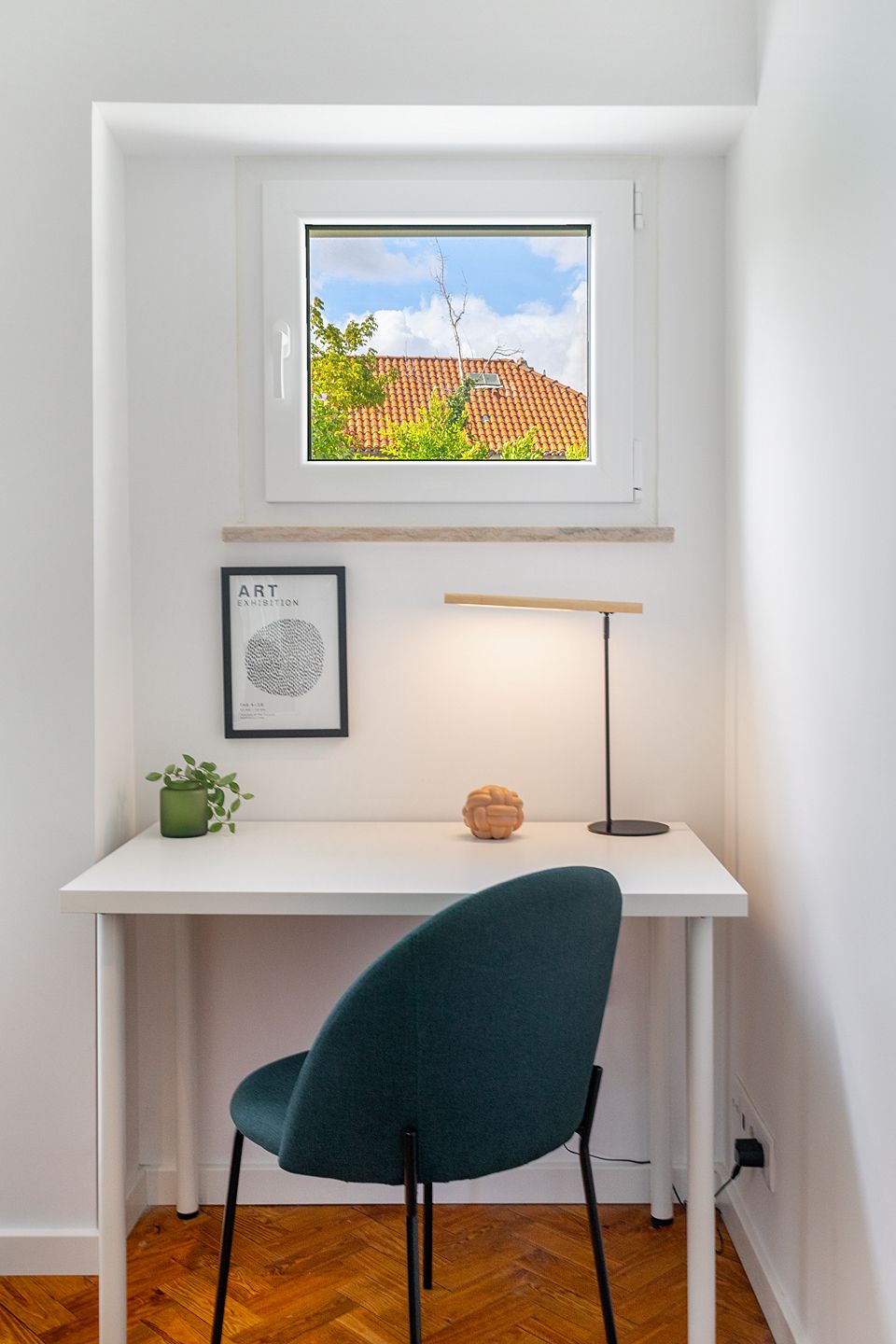 A white desk with a teal chair, a framed artwork, a small plant, and a lamp.