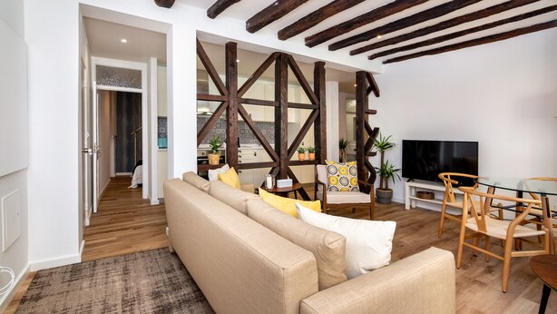 A living room with a sofa, chairs, wooden beams, a TV, and a kitchen in the background. - Boho-Chic Santos Apartment: Industriais 7 (Lisbon)