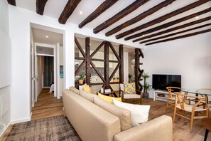 Una sala de estar con sofá, sillas, vigas de madera, televisor y una cocina al fondo.