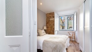 A bedroom with a bed, a chair, a window with a view of buildings, and a stone wall. - Boho-Chic Santos Apartment: Industriais 7 (Lisbon)