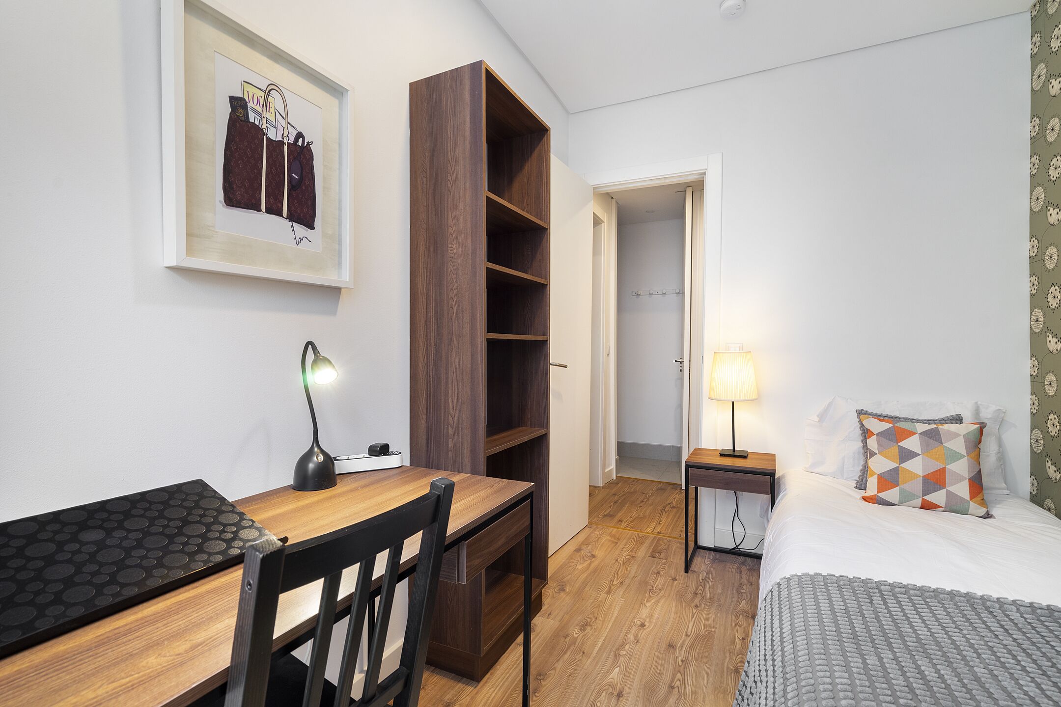 Ein gemütlicher Raum mit einem Holztisch, einem Stuhl, einem Bett mit einem gemusterten Kissen, einem Nachttisch mit einer Lampe und einem Kleiderschrank.
