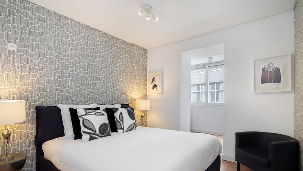 A neatly arranged bedroom with a patterned wall, a bed with black and white bedding, two bedside lamps, a black chair, and a framed picture on the wall.