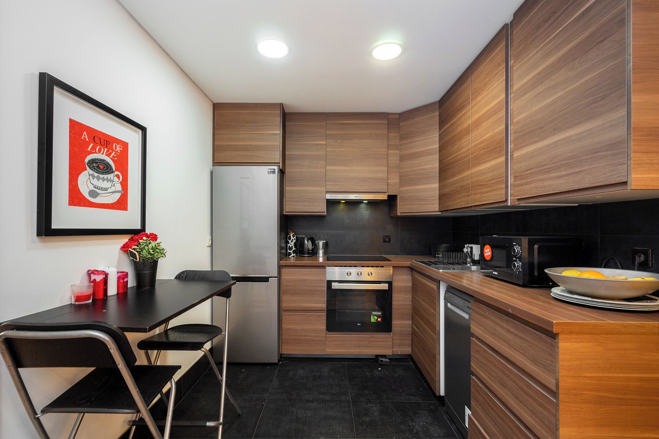 A modern kitchen with wooden cabinets, a stainless steel refrigerator, an oven, a microwave, and a dining area with a table and chairs.