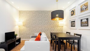 A modern living room with a white sofa, a dining table with chairs, a TV on a stand, and a patterned wallpaper.