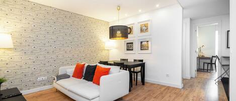 A modern living room with a white sofa, wooden flooring, a patterned wall, and a dining area with a table and chairs.