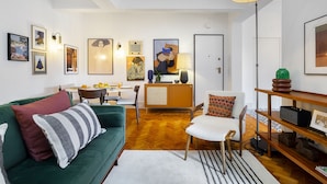 A modern living room with a green sofa, a white armchair, and a wooden coffee table. There are framed artworks on the walls and a lamp hanging from the ceiling. - Premium Alfama 2-Br Great For Remote Workers & Digital Nomads (Lisbon)