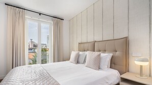 A modern hotel room with a large bed, a bedside table with a lamp, and a view of the city through large windows.