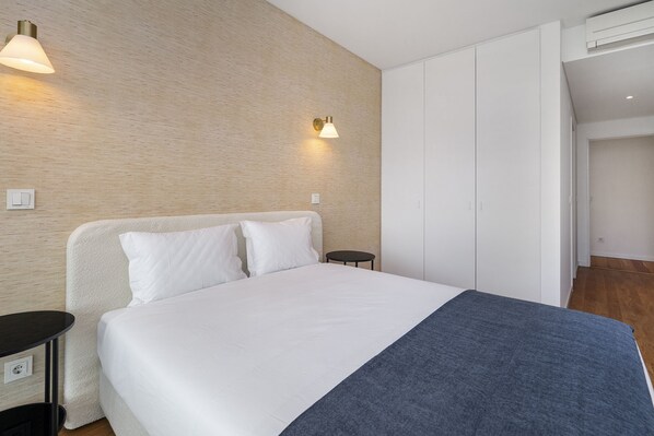Una cama bien hecha con sábanas blancas y un plaid azul, una mesita de noche pequeña, una lámpara de pared y un armario con puertas correderas.