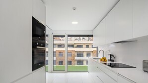 A modern kitchen with white cabinets, a black induction cooktop, and a view of a residential building through a glass door. - Urban Retreat W/Parking, Elevator & Ac By Benfica Stadium (Lisbon)
