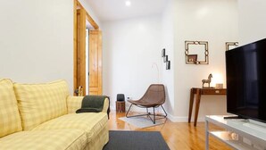 Una sala de estar con un sofá amarillo a cuadros, una silla marrón, un televisor sobre un mueble blanco y una puerta de madera que da a otra habitación.