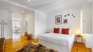 A modern bedroom with a large bed, wooden flooring, a fur rug, and a dining area visible in the background.