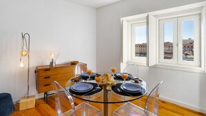A dining area with a round glass table set for four, a wooden sideboard, and a city view through large windows.
