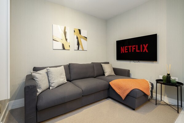 A modern living room with a grey sectional sofa, a flat-screen TV mounted on the wall, abstract art pieces, and a small side table with decorative items. - Lisbon'S Best Kept Secret: 2-Bed W/Ac & Patio (Lisbon)