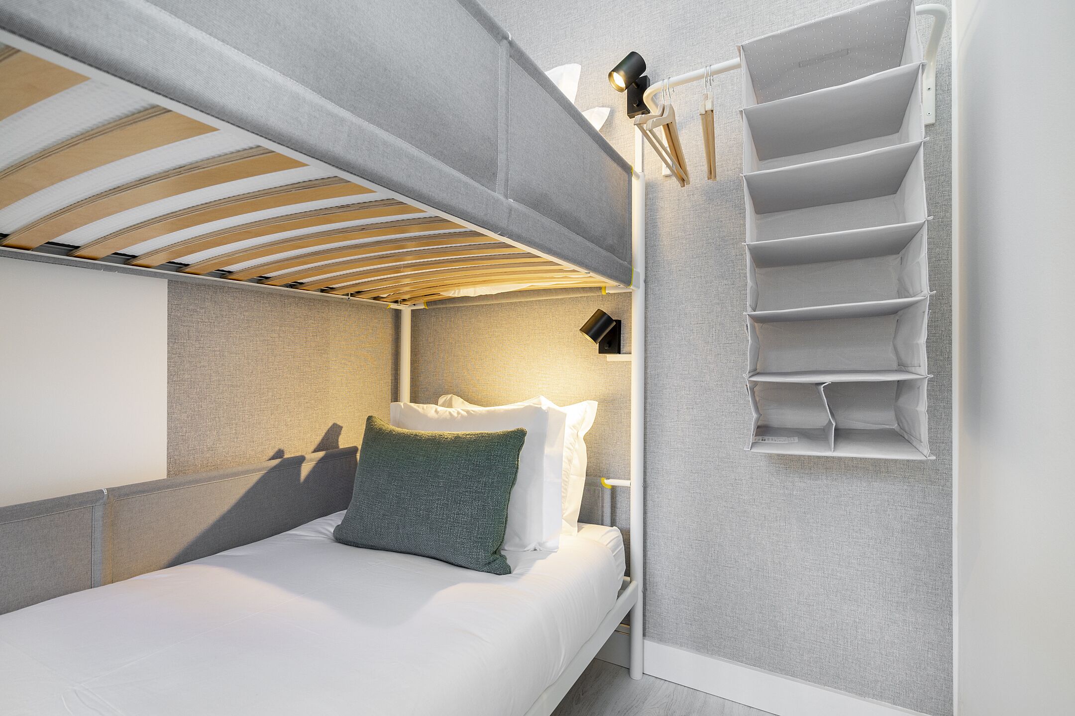 Ein modernes Hotelzimmer mit Etagenbett, Nachttisch mit Lampe und einem an der Wand befestigten Stauraum.