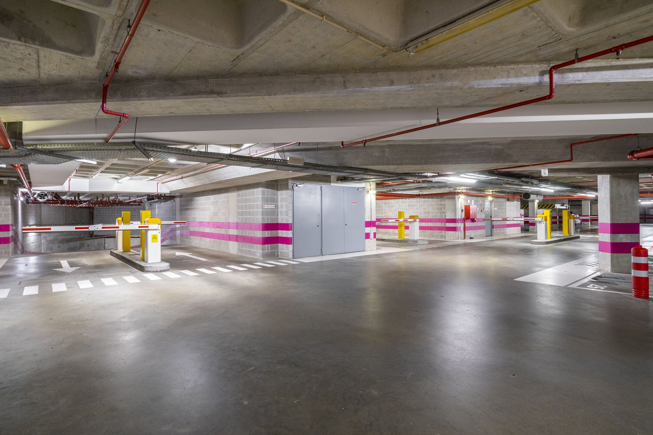 Un estacionamiento subterráneo con pilares de hormigón, barreras de seguridad amarillas y rojas, y flechas direccionales en el suelo.