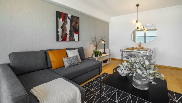 En moderne stue med en grå sofa, et sofabord med en vase, og et spiseområde med et spejl og et bord.
