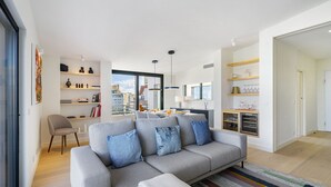 Un salon moderne avec un canapé gris, des coussins bleus et un tapis à rayures. On y trouve un coin repas avec une table et des chaises, ainsi qu'une cuisine avec des meubles blancs et une fenêtre.