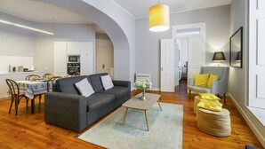 Una sala de estar moderna con un sofá gris, una mesa de centro y otomanas. Hay un comedor con una mesa y sillas, y una cocina al fondo.